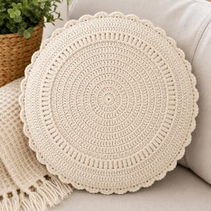 May include: A round, cream-coloured crochet cushion featuring a dense, concentric circular pattern with a scalloped edge. The texture is created by intricate yarn stitching, forming a uniform, radial design that radiates from the centre outwards.
