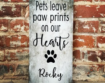 May include: A rectangular wooden sign with a distressed gray background and black text. The sign reads "Pets leave paw prints on our Hearts" with a paw print graphic and the name "Rocky" below. The sign is propped against a brick wall.