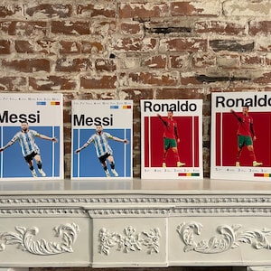 May include: Four framed prints of soccer players, two of Lionel Messi and two of Cristiano Ronaldo. The prints are on a white mantelpiece with ornate detailing. The prints feature the players in their national team jerseys, with the text "Messi" and "Ronaldo" in bold black lettering.