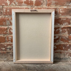 May include: The back of a wooden framed canvas with a light beige canvas stretched over it. The frame is a light wood color and has a metal hanging hook on the top.
