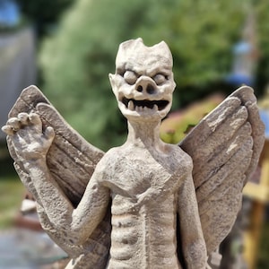 May include: A beige stone gargoyle statue featuring a winged creature with a textured, weathered surface. The figure has a pointed head, wide eyes, a flat nose, and an open mouth revealing sharp teeth. It stands with one arm raised and wings spread.
