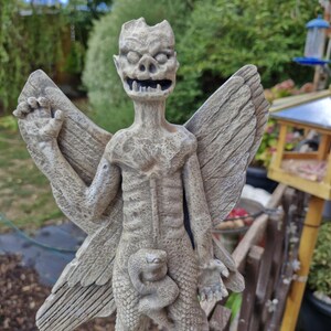 May include: A light gray stone-colored garden statue of a winged creature with a textured, weathered surface. The figure has a pointed face, bared teeth, and large wings extending from its back. A small snake-like form is coiled around its midsection.