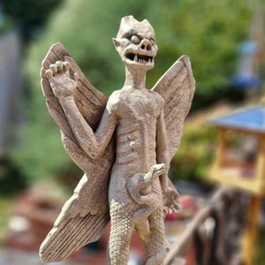 May include: A beige stone-colored statue of a winged creature with pointed ears, bared teeth, and a textured body. The figure has large, detailed wings extending from its back and a snake coiled around its lower torso, with the snake head visible.