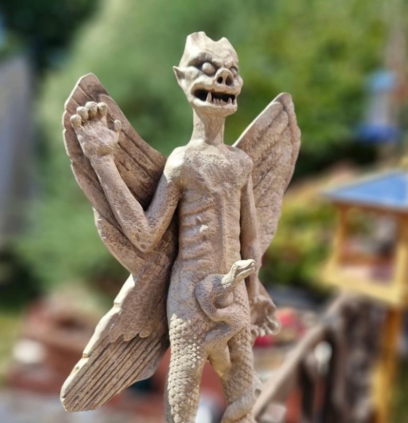 May include: A beige stone-colored statue of a winged creature with pointed ears, bared teeth, and a textured body. The figure has large, detailed wings extending from its back and a snake coiled around its lower torso, with the snake head visible.