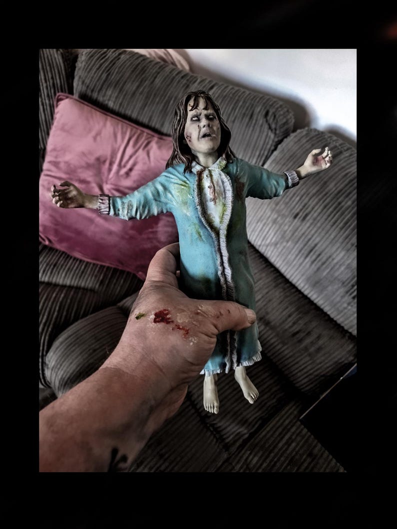 May include: A detailed figurine of a person with pale skin, dark hair, and white eyes, wearing a long-sleeved, light blue nightgown with a ruffled collar and cuffs. The figure has a distressed appearance with dark smudges and stains on the fabric.