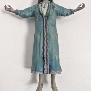 May include: A figurine of a person with pale, grayish skin and dark hair, wearing a long-sleeved, light blue robe with ruffled trim. The figure has arms outstretched to the sides, a distressed facial expression, and visible stains on the robe.
