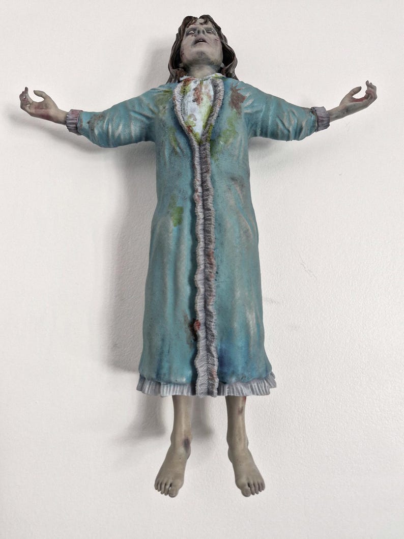 May include: A figurine of a person with pale, grayish skin and dark hair, wearing a long-sleeved, light blue robe with ruffled trim. The figure has arms outstretched to the sides, a distressed facial expression, and visible stains on the robe.