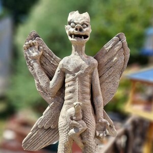 May include: A beige stone-colored statue of a winged creature with a textured, scaly body, pointed ears, and bared teeth. The figure has large, detailed wings extending from its back and holds a smaller, indistinct creature against its midsection.