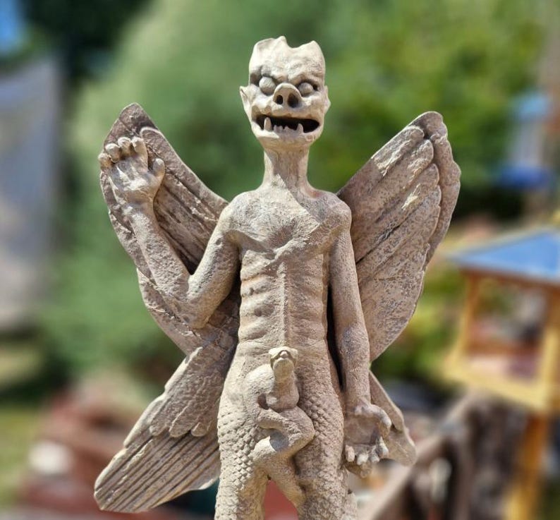 May include: A beige stone-colored statue of a winged creature with a textured, scaly body, pointed ears, and bared teeth. The figure has large, detailed wings extending from its back and holds a smaller, indistinct creature against its midsection.