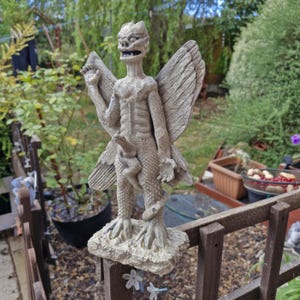 May include: A light gray stone-colored garden statue depicting a winged creature with a textured, scaly body, pointed ears, and an open mouth. The figure stands on a rectangular base, featuring large feathered wings and a snake coiled around its legs.