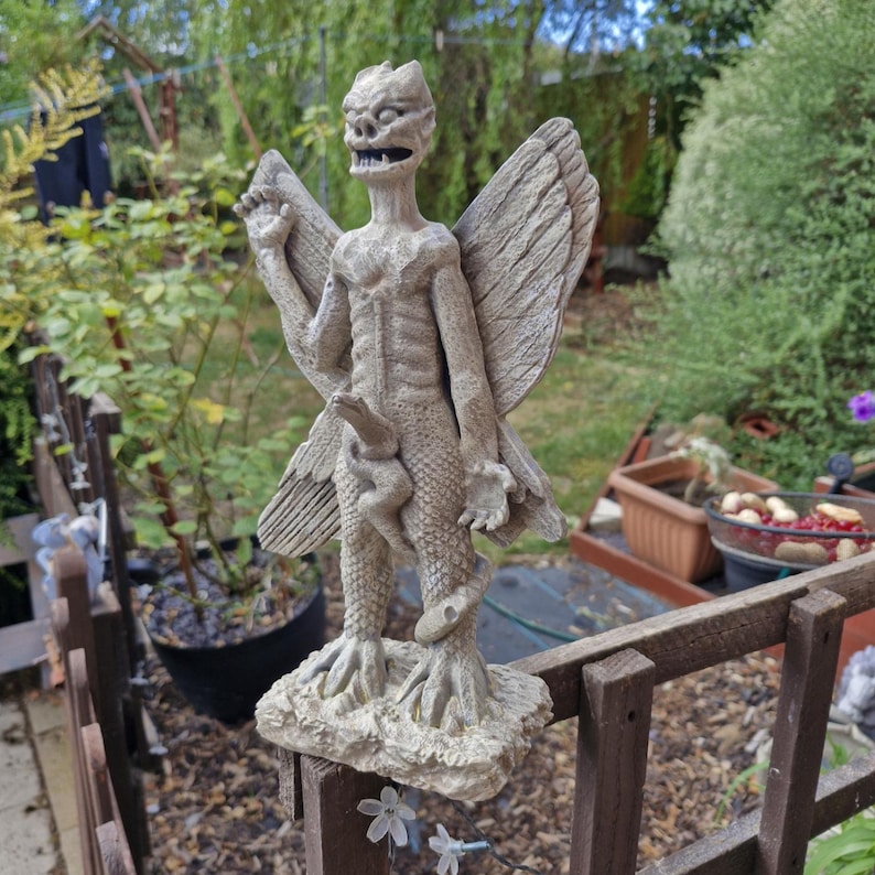 May include: A light gray stone-colored garden statue depicting a winged creature with a textured, scaly body, pointed ears, and an open mouth. The figure stands on a rectangular base, featuring large feathered wings and a snake coiled around its legs.