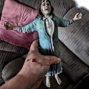 May include: A detailed figurine of a character with disheveled brown hair, pale skin, and white eyes, wearing a light blue nightgown with ruffled trim and dark stains. The figure has arms outstretched and is depicted with a distressed facial expression.