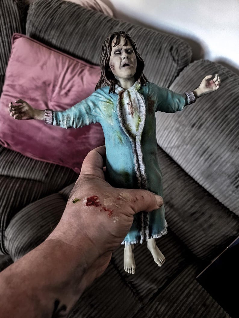 May include: A detailed figurine of a character with disheveled brown hair, pale skin, and white eyes, wearing a light blue nightgown with ruffled trim and dark stains. The figure has arms outstretched and is depicted with a distressed facial expression.