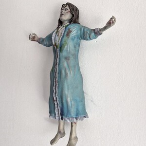 May include: A figurine of a person with long brown hair, wearing a light blue nightgown with ruffled trim and dark stains. The figure is posed with arms outstretched and head tilted back, featuring pale skin tones and a distressed, weathered appearance.