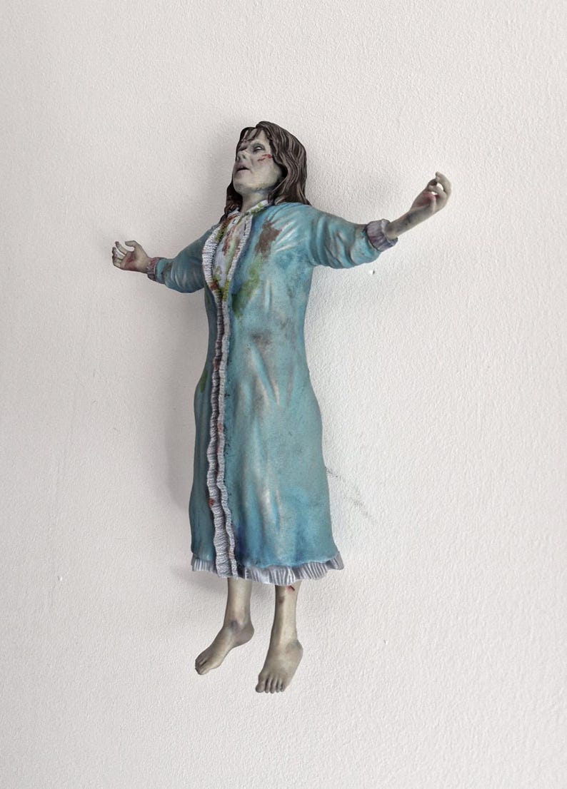 May include: A figurine of a person with long brown hair, wearing a light blue nightgown with ruffled trim and dark stains. The figure is posed with arms outstretched and head tilted back, featuring pale skin tones and a distressed, weathered appearance.
