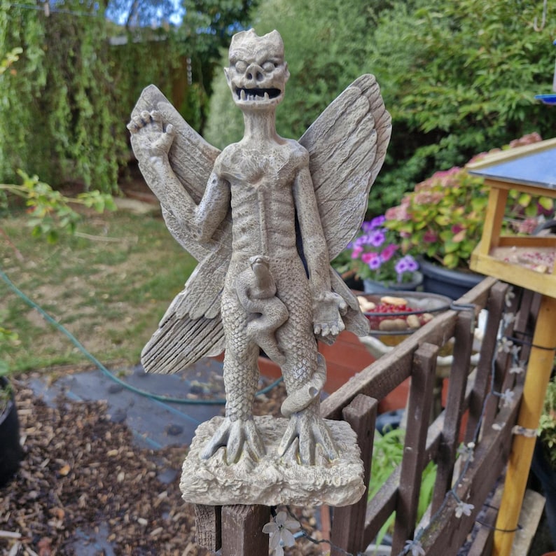 May include: A stone-colored statue of a winged creature with a horned head, sharp teeth, and a textured, scaly body. The figure stands on a rectangular base, with one arm raised and a snake coiled around its midsection, featuring detailed wings.