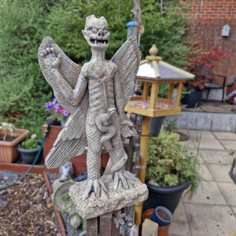 May include: A stone-colored garden statue of a winged creature with pointed ears, sharp teeth, and a textured body. The figure stands on a rectangular base, featuring a snake coiled around its legs and one arm raised with an open palm.