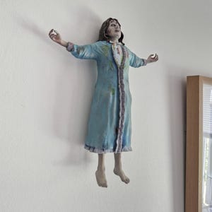 May include: A wall-mounted figurine depicting a figure with long dark hair, wearing a light blue nightgown with ruffled trim and stained details. The figure is posed with arms outstretched and head tilted back, featuring pale skin and dark eye markings.