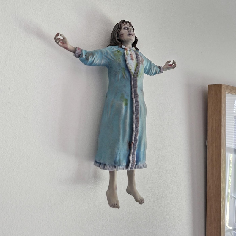 May include: A wall-mounted figurine depicting a figure with long dark hair, wearing a light blue nightgown with ruffled trim and stained details. The figure is posed with arms outstretched and head tilted back, featuring pale skin and dark eye markings.