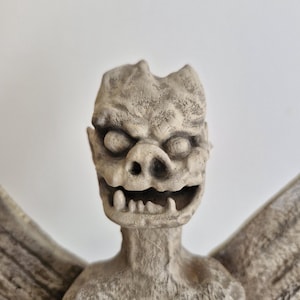 May include: A stone-colored gargoyle figurine featuring a textured, weathered surface, prominent pointed ears, deep-set circular eyes, a flat nose, and an open mouth displaying a row of sharp, jagged teeth, with wings extending from the sides.