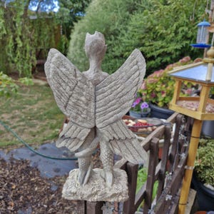 May include: A gray stone-colored garden statue of a winged creature with a textured, scaly tail and large, detailed feathered wings. The figure stands on a rectangular base, viewed from the back, showcasing intricate wing plumage and a slender form.