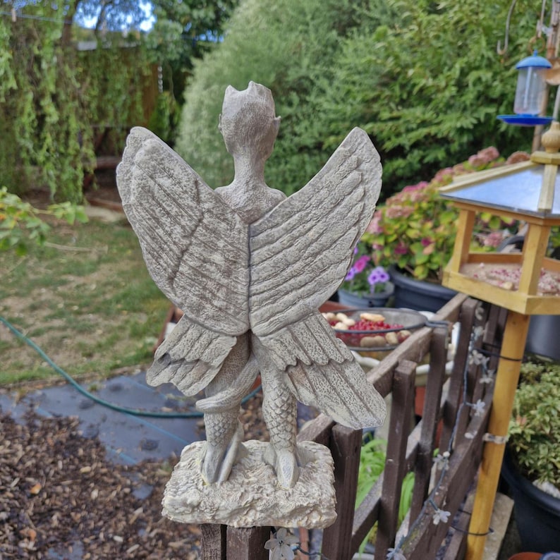 May include: A gray stone-colored garden statue of a winged creature with a textured, scaly tail and large, detailed feathered wings. The figure stands on a rectangular base, viewed from the back, showcasing intricate wing plumage and a slender form.