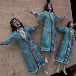 May include: Three identical figurines of a girl with dark hair, wearing a teal nightgown with a ruffled white front panel and sleeves. Each figure is posed with arms outstretched, featuring distressed fabric details and a pale, expressive facial expression.