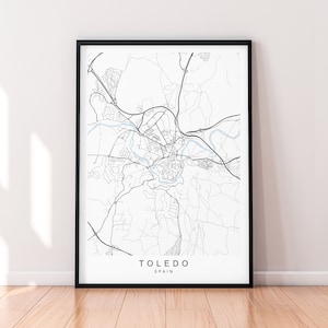 Op de afbeelding: Een ingelijste zwart-wit kaartprint van Toledo, Spanje. De kaart toont gedetailleerde straatindelingen, wegen en een rivier. De naam van de stad en het land zijn in een strak, modern lettertype onderaan de print gedrukt.