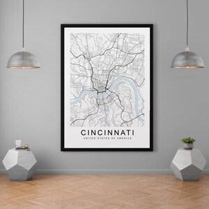 Może przedstawiać: Czarno-biały wydruk mapy Cincinnati, Ohio, Stany Zjednoczone Ameryki, w czarnej ramie. Mapa przedstawia ulice i drogi wodne miasta. Wydruk wisi na szarej ścianie nad drewnianą podłogą.