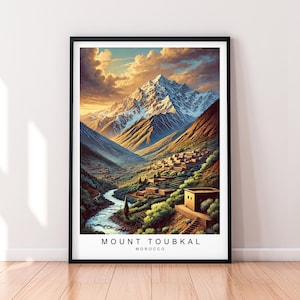 Op de afbeelding: Een ingelijste print van een berglandschap met een dorp in de vallei. De berg is bedekt met sneeuw en de lucht is een levendig oranje en geel. De tekst "MOUNT TOUBKAL MOROCCO" staat onderaan de print.
