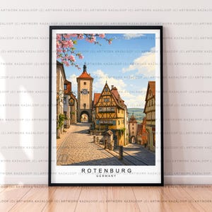 Può includere: Una stampa artistica incorniciata raffigurante una strada acciottolata a Rotenburg, in Germania, con edifici a graticcio, una torre dell'orologio e fiori di ciliegio rosa in primo piano. Il testo ROTENBURG GERMANY è stampato in nero in basso.