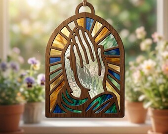 May include: A wooden hanging ornament featuring an arched frame with a cutout design of praying hands. The interior is filled with textured, translucent panels in shades of blue, green, and amber, creating a stained-glass effect when light shines through.