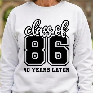 May include: A white long-sleeve sweatshirt featuring a black graphic print on the chest. The text reads "class of 86" in a stylized script font above large block numbers "86" and the text "40 YEARS LATER" in a bold serif font below.