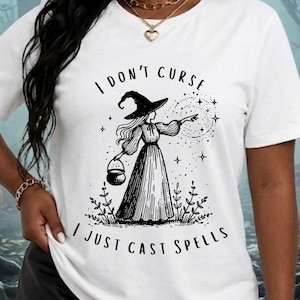 Könnte beinhalten: Ein weißes T-Shirt mit einer schwarzen grafischen Illustration einer Hexe mit spitzem Hut und langem Kleid, die einen kleinen Kessel hält. Der Text "I DON'T CURSE I JUST CAST SPELLS" ist in schwarzer Schrift über und unter der Figur gedruckt.