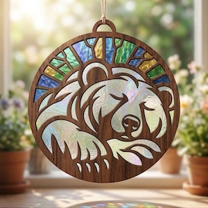 May include: A circular wooden hanging ornament featuring a laser-cut design of a sleeping bear. The bear's body is filled with iridescent, pearlescent glass, while the upper border displays a sunburst pattern of blue, green, and yellow glass.
