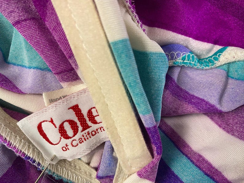 May include: Close-up of a vintage garment with a striped pattern in shades of purple, teal, and white. The label reads "Cole of California" in red lettering. The fabric is soft and appears to be a lightweight material.
