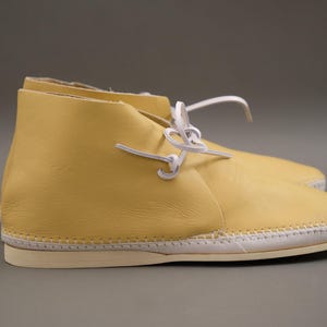 Pu&ograve; includere: Un paio di stivaletti in pelle gialla con lacci bianchi. Le scarpe hanno una suola cucita e una punta arrotondata. Gli stivaletti sono di colore giallo chiaro e sembrano nuovi.