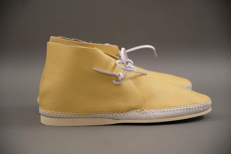 Pu&ograve; includere: Un paio di stivaletti in pelle gialla con lacci bianchi. Le scarpe hanno una suola cucita e una punta arrotondata. Gli stivaletti sono di colore giallo chiaro e sembrano nuovi.