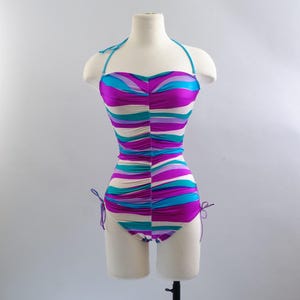 May include: A vintage one-piece swimsuit with a halter neck and a vibrant, wavy pattern in shades of purple, teal, and cream. The swimsuit is ruched and has adjustable ties at the sides. The garment is displayed on a mannequin.