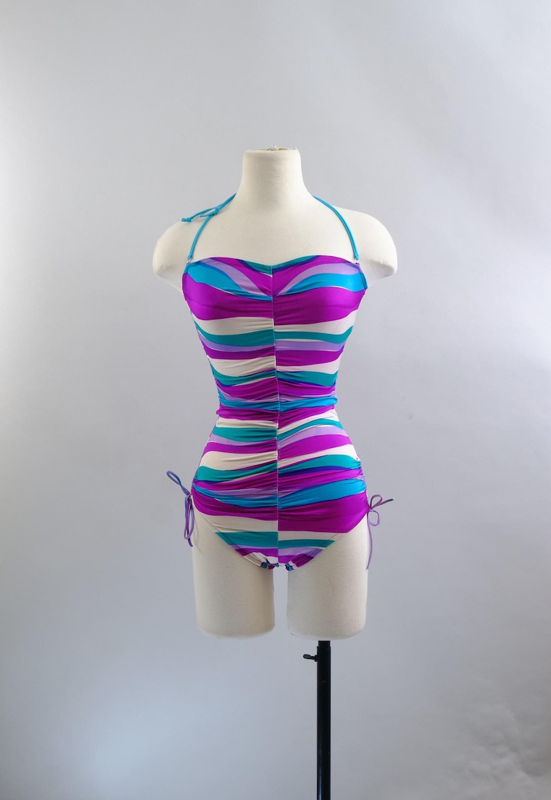 May include: A vintage one-piece swimsuit with a halter neck and a vibrant, wavy pattern in shades of purple, teal, and cream. The swimsuit is ruched and has adjustable ties at the sides. The garment is displayed on a mannequin.