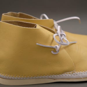 Pu&ograve; includere: Un paio di stivaletti chukka in pelle gialla con lacci bianchi. Gli stivali hanno una suola cucita. Le scarpe sono di colore giallo chiaro. Lo sfondo &egrave; neutro.
