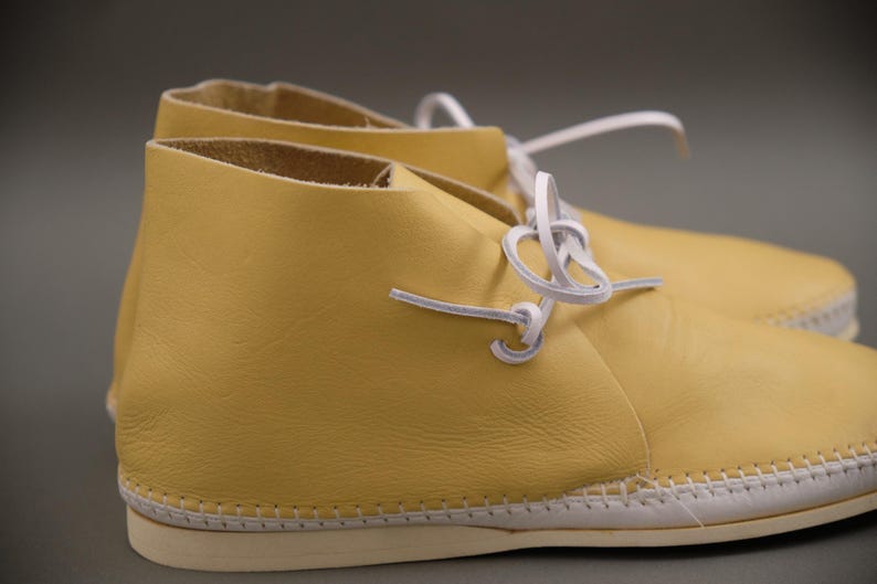 Pu&ograve; includere: Un paio di stivaletti chukka in pelle gialla con lacci bianchi. Gli stivali hanno una suola cucita. Le scarpe sono di colore giallo chiaro. Lo sfondo &egrave; neutro.