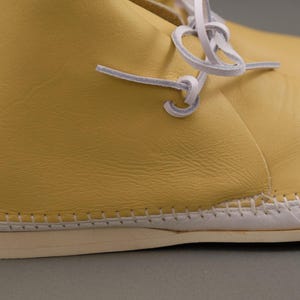 Pu&ograve; includere: Primo piano di un paio di scarpe in pelle gialla con cuciture e lacci bianchi. Le scarpe hanno una suola colorata chiara e un aspetto morbido e strutturato. I lacci sono annodati in un nodo semplice.