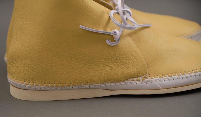 Pu&ograve; includere: Primo piano di un paio di scarpe in pelle gialla con cuciture e lacci bianchi. Le scarpe hanno una suola colorata chiara e un aspetto morbido e strutturato. I lacci sono annodati in un nodo semplice.