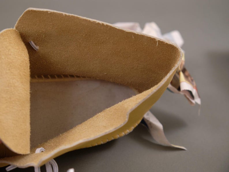 Pu&ograve; includere: Primo piano di un astuccio in pelle beige con un interno morbido e strutturato. I bordi sono ripiegati, rivelando l'interno dell'astuccio. &Egrave; visibile un filo bianco. Lo sfondo &egrave; grigio neutro.