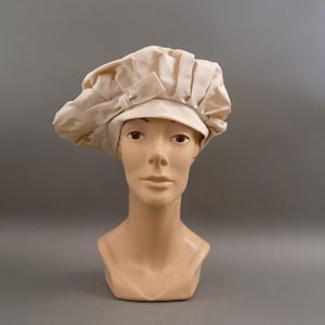May include: A cream-coloured chef's hat with a brim and a bow detail, displayed on a mannequin head. The hat has a pleated design and a soft, textured appearance. The mannequin is a neutral skin tone.
