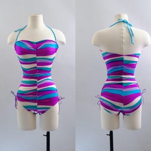 May include: A vintage-style swimsuit with a halter top, displayed on a mannequin. The one-piece suit features horizontal stripes in shades of purple, teal, and white. The suit has tie details on the sides and a matching teal halter strap.