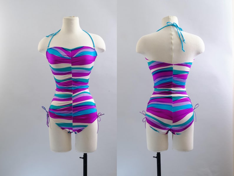 May include: A vintage-style swimsuit with a halter top, displayed on a mannequin. The one-piece suit features horizontal stripes in shades of purple, teal, and white. The suit has tie details on the sides and a matching teal halter strap.