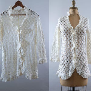 May include: Off-white, long-sleeved lace top with a ruffled collar and front. The top features a button-up design and a scalloped hem. The lace has a circular pattern. The garment is displayed on a wooden hanger and a mannequin.
