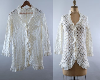 May include: Off-white, long-sleeved lace top with a ruffled collar and front. The top features a button-up design and a scalloped hem. The lace has a circular pattern. The garment is displayed on a wooden hanger and a mannequin.